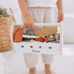 May include: A white wooden toy toolbox with a light green handle, featuring the name Luca printed in black script on the front. The box contains various wooden toy tools including a hammer, a saw, and screws in shades of orange, grey, and tan.