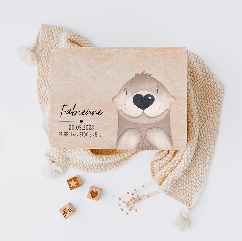 Puede incluir: Una placa rectangular de madera con una ilustraci&oacute;n impresa de una nutria de color marr&oacute;n claro con una nariz en forma de coraz&oacute;n. El texto dice Fabienne, 26.05.2023, 21:58 Uhr - 3190 g - 51 cm. La placa est&aacute; sobre una tela color crema.