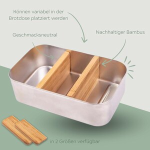 May include: A silver metal lunchbox with two removable bamboo dividers. The dividers can be placed in the lunchbox in different configurations. The lunchbox is labeled "Geschmacksneutral" and "Nachhaltiger Bambus". The text "in 2 Gr&ouml;&szlig;en verf&uuml;gbar" is also visible.