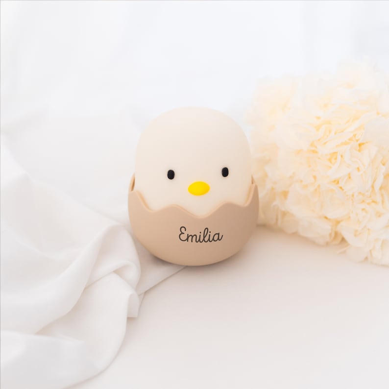May include: A smooth, rounded lamp shaped like a chick hatching from a beige eggshell. The chick has a matte white finish, two small black eyes, and a bright yellow beak. The beige eggshell base features the name Emilia in black script.