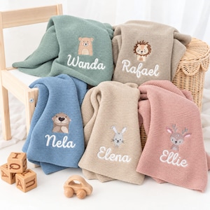Può includere: Cinque coperte lavorate a maglia per neonati in verde salvia, beige, blu, marrone chiaro e rosa polvere. Ogni coperta presenta un animale ricamato e un nome: Wanda con un orso, Rafael con un leone, Nela con una lontra, Elena con un coniglio ed Ellie con un cervo.