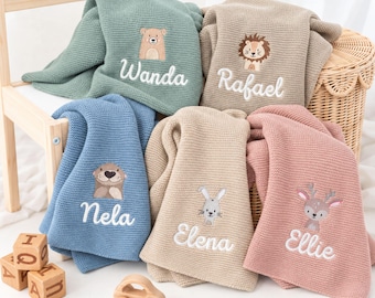 May include: Five knitted baby blankets in sage green, beige, blue, light tan, and dusty pink. Each blanket features an embroidered animal character and a name: Wanda with a bear, Rafael with a lion, Nela with an otter, Elena with a rabbit, and Ellie with a deer.