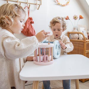May include: Two children play with a wooden toy activity center. The center has a pink and blue color scheme and features a spinning wheel with shapes, a bead maze, and a rolling ball track.