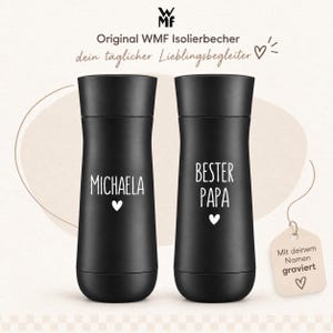 May include: Two matte black insulated travel mugs with a cylindrical shape and a lid. The left mug features the white text "MICHAELA" above a small heart icon. The right mug features the white text "BESTER PAPA" above a small heart icon.