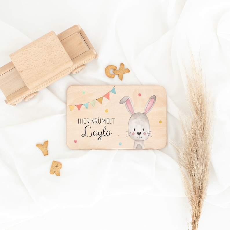 May include: A rectangular wooden board featuring a printed illustration of a gray rabbit, colorful bunting flags, and the text "HIER KR&Uuml;MELT Layla". The board is surrounded by a wooden toy truck, letter-shaped cookies, and a dried grass stalk.