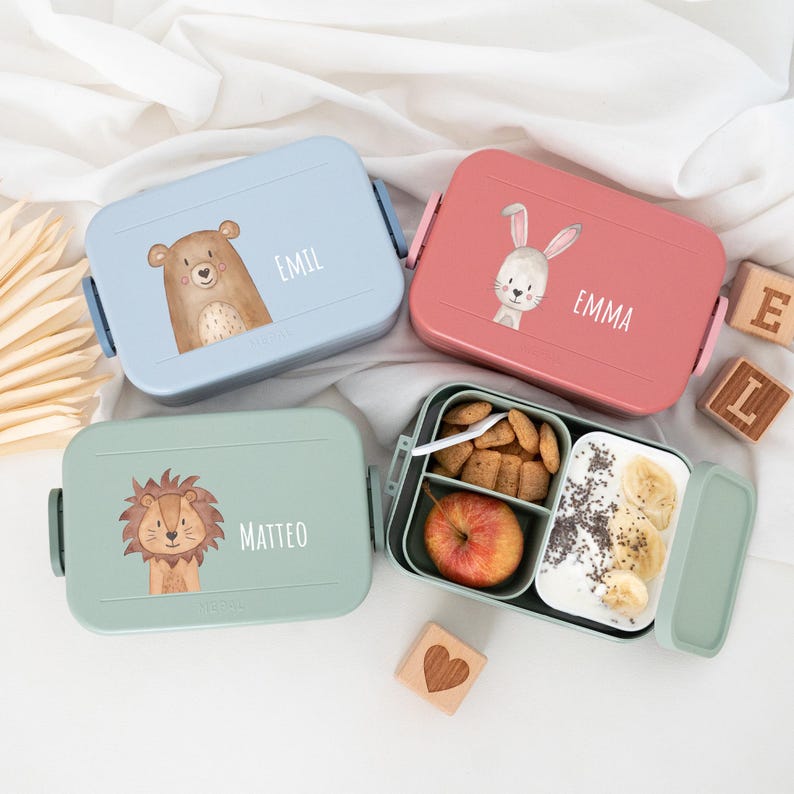 May include: Three colorful lunchboxes with animal illustrations and personalized names. The lunchboxes are blue, pink, and green. The blue lunchbox has a bear, the pink lunchbox has a rabbit, and the green lunchbox has a lion. The names on the lunchboxes are Emil, Emma, and Matteo.
