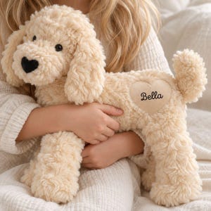 Pode incluir: Um cão de peluche feito de pelo sintético encaracolado de cor creme. O brinquedo apresenta um nariz preto em forma de coração, olhos pretos e um remendo castanho claro em forma de coração no lado com o nome Bella bordado em cursivo.