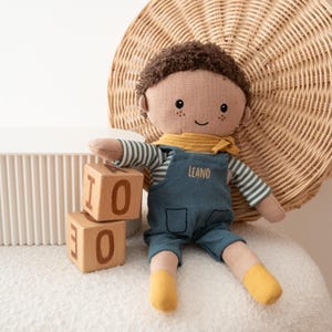 May include: A soft doll with brown curly hair, freckles, and a smiling face. The doll wears a striped shirt, blue overalls with the name "LEANO", and a yellow scarf and socks. Two wooden blocks with the numbers "10" and "0" are nearby.