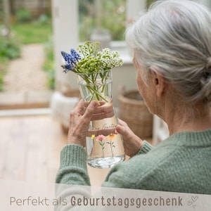 May include: A clear glass vase with a flared top, holding a bouquet of fresh flowers. The vase has a floral design and the text "Omas Garten". The bottom of the image has the text "Perfekt als Geburtstagsgeschenk".