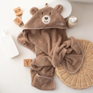May include: A brown hooded baby towel with bear ears and a smiling bear face embroidered on the hood. The towel has the name "Carlo" embroidered on it. The towel is made of soft, absorbent material.