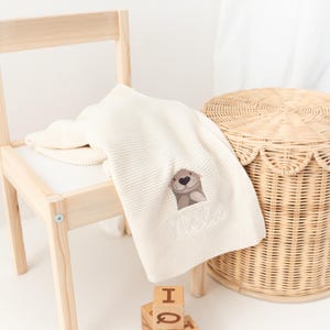 Puede incluir: Manta de beb&eacute; color crema con un dise&ntilde;o de nutria bordada y el nombre "Meka". La manta est&aacute; sobre una silla de madera. Una cesta de mimbre y bloques de madera tambi&eacute;n est&aacute;n en la imagen.