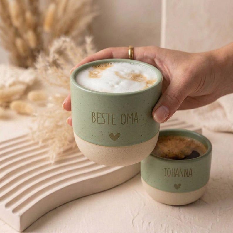 Pode incluir: Duas canecas de cer&acirc;mica com um design bicolor, apresentando uma sec&ccedil;&atilde;o superior em verde s&aacute;lvia mate e uma base em cor creme. A caneca maior tem impresso o texto BESTE OMA e um pequeno cora&ccedil;&atilde;o, enquanto a menor apresenta o nome JOHANNA e um cora&ccedil;&atilde;o.