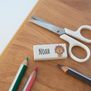 May include: A rectangular white eraser featuring the name NOAH in black text next to a small illustration of a lion's face. The eraser is positioned near a pair of white-handled scissors and two coloured pencils, one green and one pink.