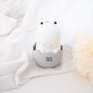 May include: A white dinosaur-shaped night light with black eyes and a small green detail on its head, sitting inside a light gray egg-shaped base. The base features the name THEO printed in black, sans-serif capital letters on the front.