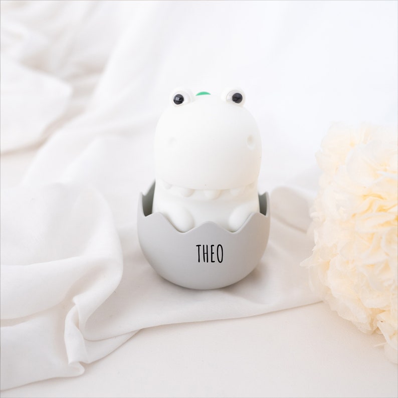 May include: A white dinosaur-shaped night light with black eyes and a small green detail on its head, sitting inside a light gray egg-shaped base. The base features the name THEO printed in black, sans-serif capital letters on the front.
