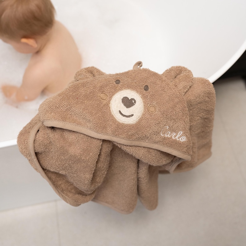 May include: A brown hooded baby towel with a bear face design. The towel has a light brown trim and the name "Carlo" embroidered on the hood. The towel is made of soft material and is designed for use after bath time.