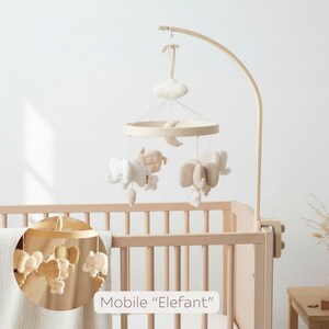 May include: A wooden baby mobile with hanging plush elephants, a moon, and a cloud. The mobile is suspended above a wooden crib. The text "Mobile 'Elefant'" is visible.
