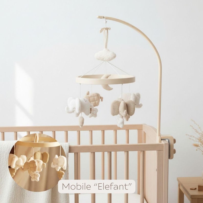 May include: A wooden baby mobile with hanging plush elephants, a moon, and a cloud. The mobile is suspended above a wooden crib. The text "Mobile 'Elefant'" is visible.