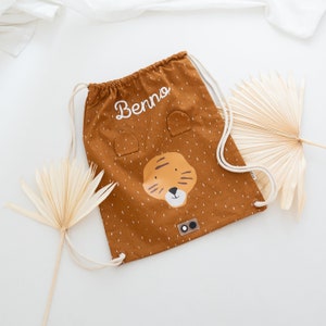 May include: A brown drawstring bag with a tiger face design and the name "Benno" embroidered on it. The bag has white drawstrings and a white polka dot pattern.