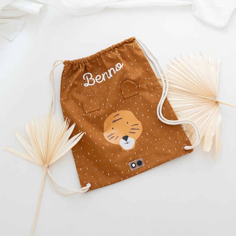 May include: A brown drawstring bag with a tiger face design and the name "Benno" embroidered on it. The bag has white drawstrings and a white polka dot pattern.