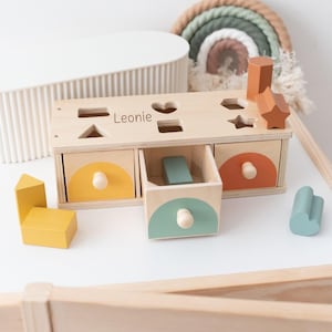 Könnte beinhalten: Holz-Formensortierspielzeug mit drei Schubladen und verschiedenen Formblöcken. Die Schubladen sind mit gelben, orangefarbenen und türkisfarbenen Halbkreisen bemalt. Der Name "Leonie" ist oben aufgedruckt. Ein Regenbogen und anderes Holzspielzeug sind im Hintergrund.