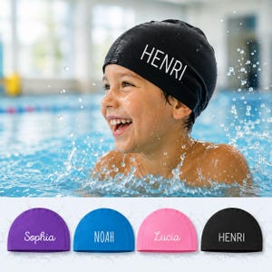 Op de afbeelding: Een zwarte zwemmuts met de naam HENRI in witte hoofdletters. De collectie bevat vier zwemmutsen in paars, blauw, roze en zwart, elk voorzien van een gepersonaliseerde naam in witte tekst, namelijk Sophia, NOAH, Lucia en de naam HENRI.