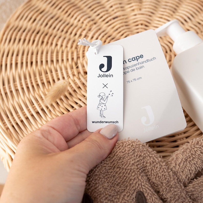 May include: A white tag with the brand name "Jollein" and a drawing of a child, attached to a brown hooded towel. The tag also includes the text "wunderwunsch". A white bottle with a pump is visible in the background.