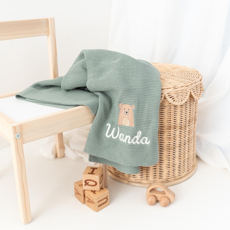 Puede incluir: Una manta de beb&eacute; de punto verde salvia con un aplique de oso y el nombre "Wanda" bordado en blanco. La manta est&aacute; sobre una peque&ntilde;a silla de madera y una cesta de mimbre. Tambi&eacute;n se ven bloques de madera y un coche de juguete.