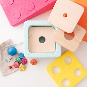 Può includere: Una collezione di giocattoli in legno colorati, tra cui un quadrato azzurro con un foro centrale, quadrati di legno con fori e un blocco rosa con rientranze circolari. È presente anche un piccolo sacchetto con il testo "Stapel Welt".