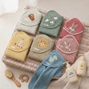 Könnte beinhalten: Eine Auswahl an Baby-Kapuzenhandtüchern in verschiedenen Farben, jeweils mit einem Tier und einem Namen bestickt. Die Handtücher sind in einem Weidenkorb angeordnet, daneben Baby-Lotion und Waschflaschen. Eine Holzbürste und ein Beißring sind ebenfalls zu sehen.