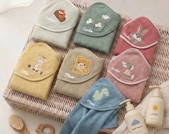May include: A collection of hooded baby towels in various colors, each embroidered with a different animal and a name. The towels are arranged in a wicker basket, with baby lotion and wash bottles nearby. A wooden brush and teether are also visible.