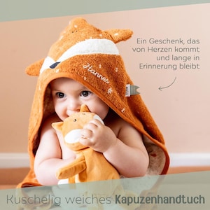 May include: A baby wearing an orange fox-themed hooded towel, holding a matching plush toy. The towel has white accents and the name "Hannes" embroidered on it. Text on the image reads "A gift that comes from the heart and remains in memory for a long time."