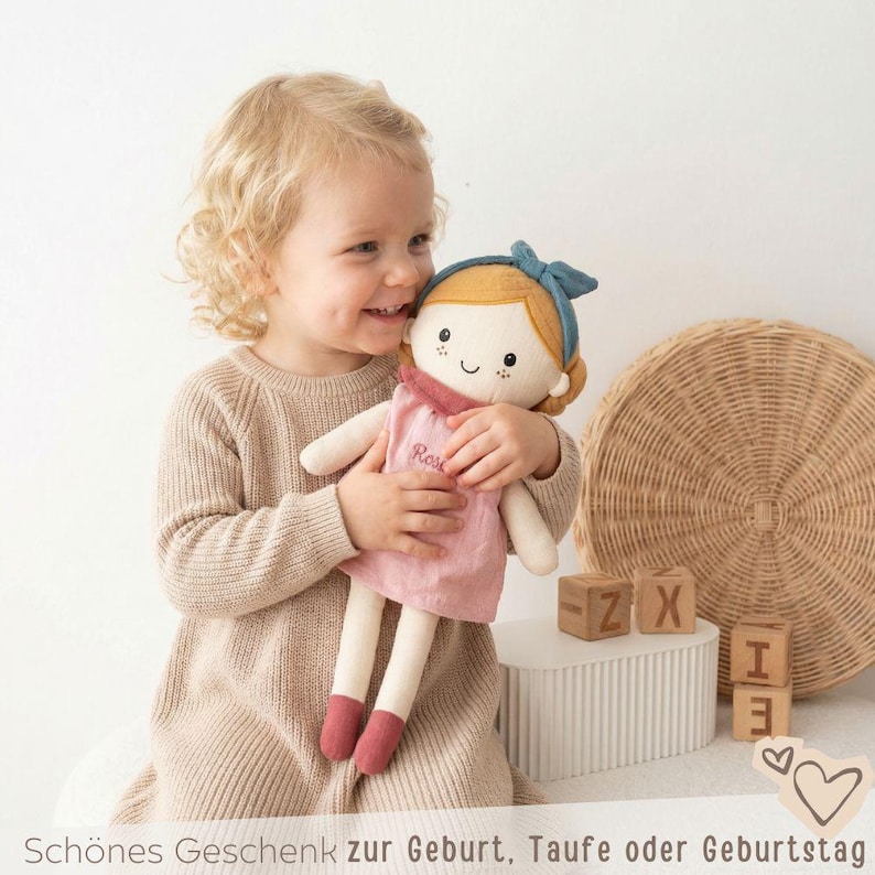 May include: A soft, handmade doll with blonde hair, a blue headband, and a pink dress. The doll is being held by a child. The doll has the name "Rose" embroidered on the dress. The image also includes wooden blocks and a woven circular decoration.