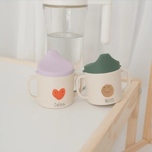 Può includere: Due tazze con beccuccio per bambini color bianco sporco con due manici. La tazza a sinistra ha un coperchio color lavanda e un cuore arancione con una faccina sorridente sopra il nome Celina. La tazza a destra ha un coperchio verde scuro e un cerchio beige con una faccina sorridente sopra il nome Matteo.
