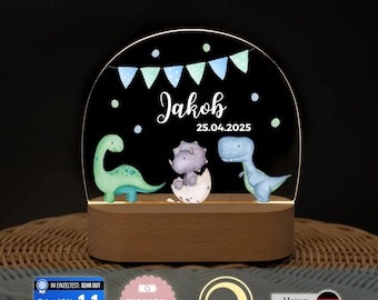 May include: An arched acrylic night light with a wooden base featuring a printed design of three dinosaurs, a bunting banner, and the text "Jakob" and "25.04.2025". The dinosaurs include a green long-necked one, a purple one in an egg, and a blue one.