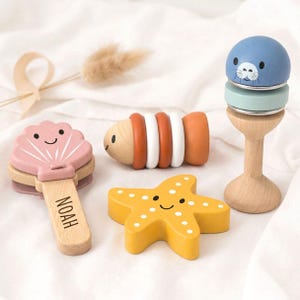 Könnte beinhalten: Ein Set aus Holzspielzeug bestehend aus einer rosa Muschelrassel mit dem eingravierten Namen NOAH auf dem Griff, einem horizontalen orange-weißen Fischspielzeug, einem gelben Seestern und einer blauen Robbenfigur auf einem Holzständer.