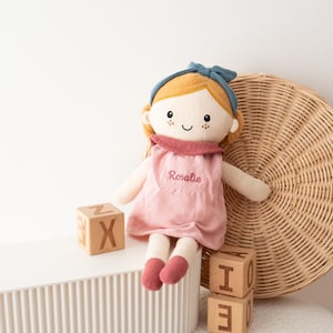 May include: A soft doll with blonde hair, a blue headband, and a pink dress embroidered with the name "Rosalie." The doll has rosy cheeks and is sitting next to wooden alphabet blocks. A woven basket is in the background.