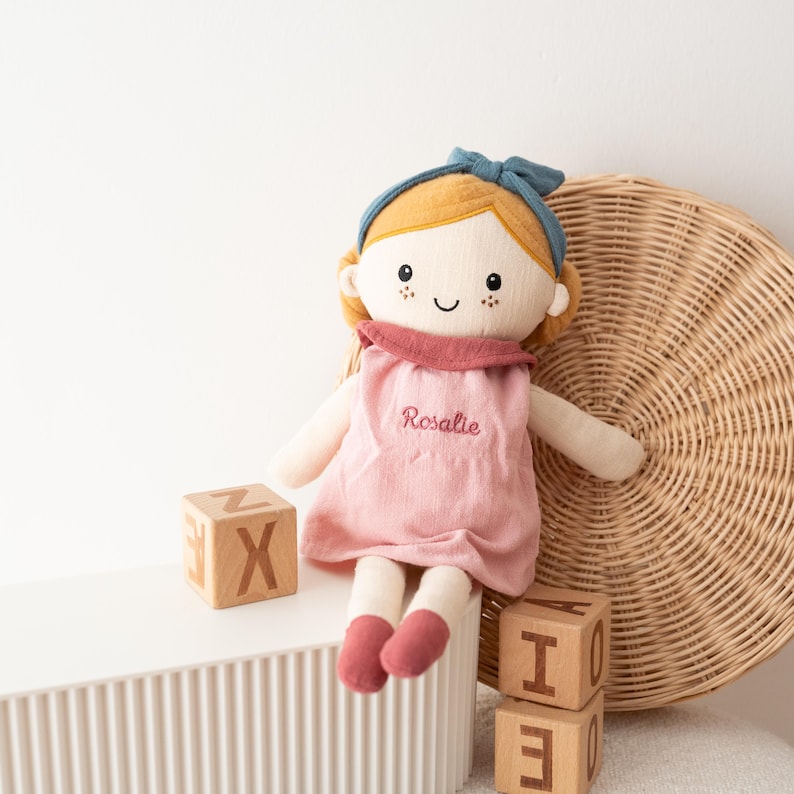 May include: A soft doll with blonde hair, a blue headband, and a pink dress embroidered with the name "Rosalie." The doll has rosy cheeks and is sitting next to wooden alphabet blocks. A woven basket is in the background.