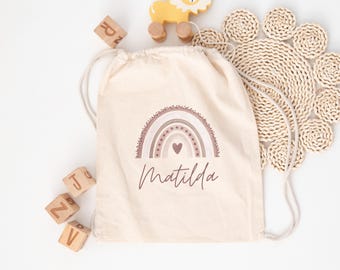 Può includere: Una borsa a cordoncino beige con un designo arcobaleno rosa e marrone e il nome Matilda stampato sotto. La borsa è circondata da blocchi di legno e un leone di legno giocattolo.
