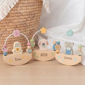 May include: Three wooden baby toys, each with a name: "Lina", "Anton", and "Emma". The toys feature a duck, a dog, and a bear. Pastel-coloured beads and floral accents adorn each toy, with a white arched handle.