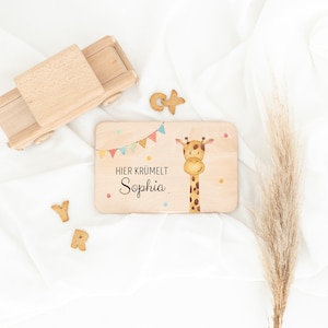 May include: A rectangular wooden board featuring a watercolor illustration of a giraffe, a colorful bunting banner, and the text "HIER KR&Uuml;MELT Sophia". The board is surrounded by a wooden toy truck, letter-shaped cookies, and a dried grass stalk.