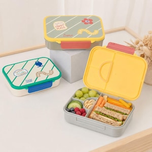 Könnte beinhalten: Drei rechteckige Bento-Lunchboxen mit Deckeln. Eine ist mit einem grünen Deckel und gelben Akzenten verschlossen, eine mit einem weißen Deckel und blauen Akzenten, und eine ist offen mit einem gelben Deckel, gefüllt mit Weintrauben, Gurken, Beeren, Crackern, Karotten und Sandwiches.