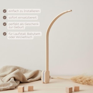 May include: A light-colored wooden baby mobile arm with a curved top. The arm is attached to a base with a gray fabric accent. The text on the image says: "easy to install", "ready to use", "perfect as a gift for birth", and "for playpen, crib or changing table."