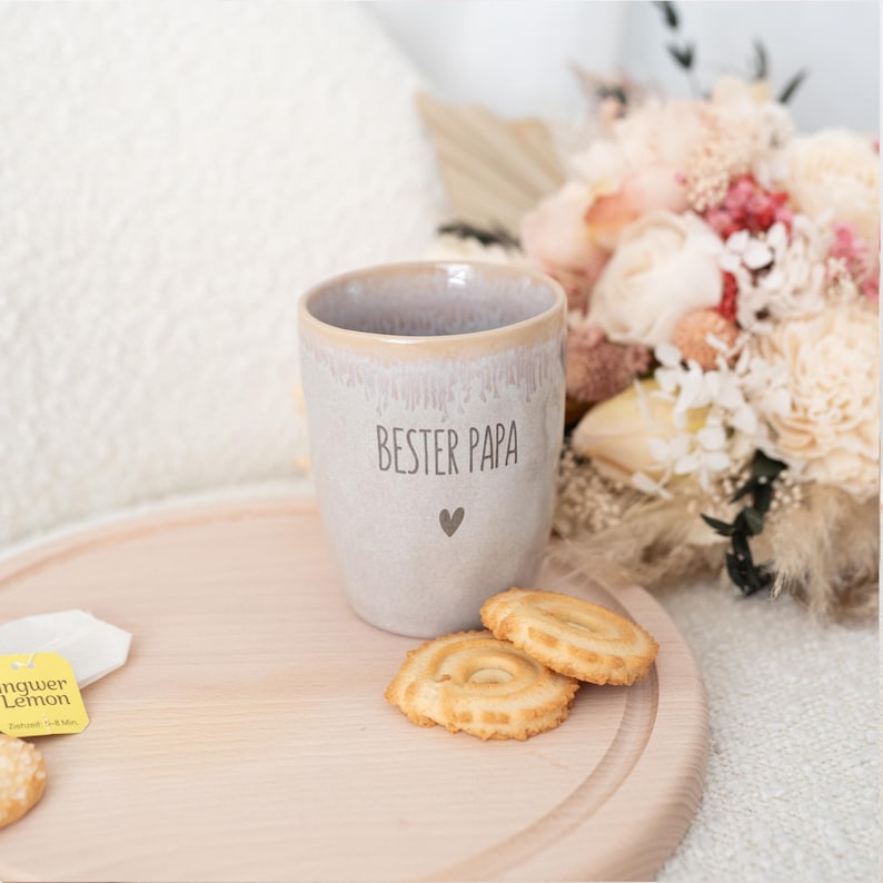 Puede incluir: Una taza de cer&aacute;mica gris claro con el texto "BESTER PAPA" y un s&iacute;mbolo de coraz&oacute;n. La taza est&aacute; sobre una bandeja de madera con galletas y una bolsita de t&eacute;. Un ramo de flores est&aacute; en el fondo. La taza tiene un borde beige.
