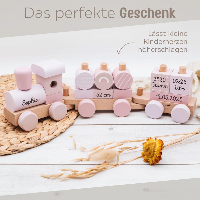 May include: A wooden toy train in pastel pink and natural wood tones. The train features the name "Sophia" on the engine, decorative blocks, and a measurement of 52 cm. Additional blocks display weight, time, and date.
