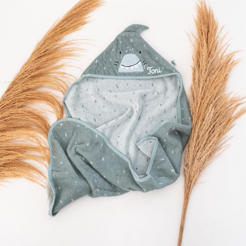 May include: A hooded baby towel in a shark design. The towel is a muted teal color with white polka dots and a shark face on the hood. The name "Toni" is embroidered on the hood. The towel is made of soft material.