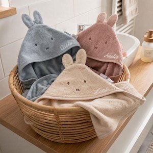 Peut inclure: Trois serviettes à capuche en bleu, vieux rose et beige, chacune dotée d'oreilles de lapin et d'un visage brodés. Les serviettes sont personnalisées avec les prénoms Carlo, Amelie et Leni cousus en fil blanc, le tout dans un panier en osier.