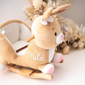 May include: A plush brown bunny rocking horse with a white bow and pink accents. The rocking horse has a white embroidered name "Mila" on the side.