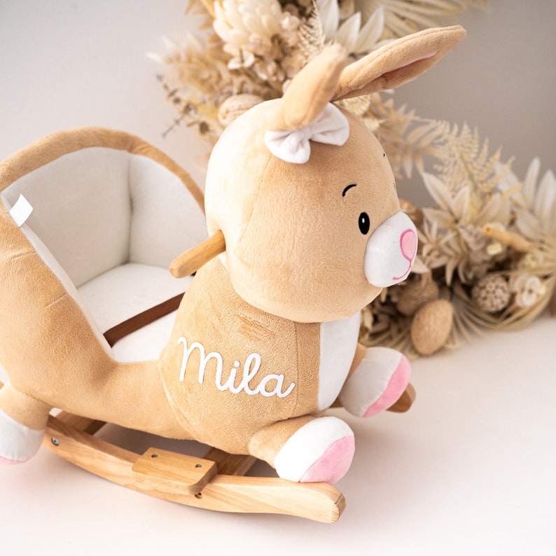May include: A plush brown bunny rocking horse with a white bow and pink accents. The rocking horse has a white embroidered name "Mila" on the side.