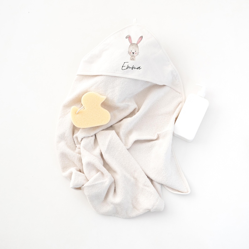 May include: A white hooded towel with a bunny illustration and the name "Emma" embroidered on the hood. A yellow rubber duck is lying on the towel.